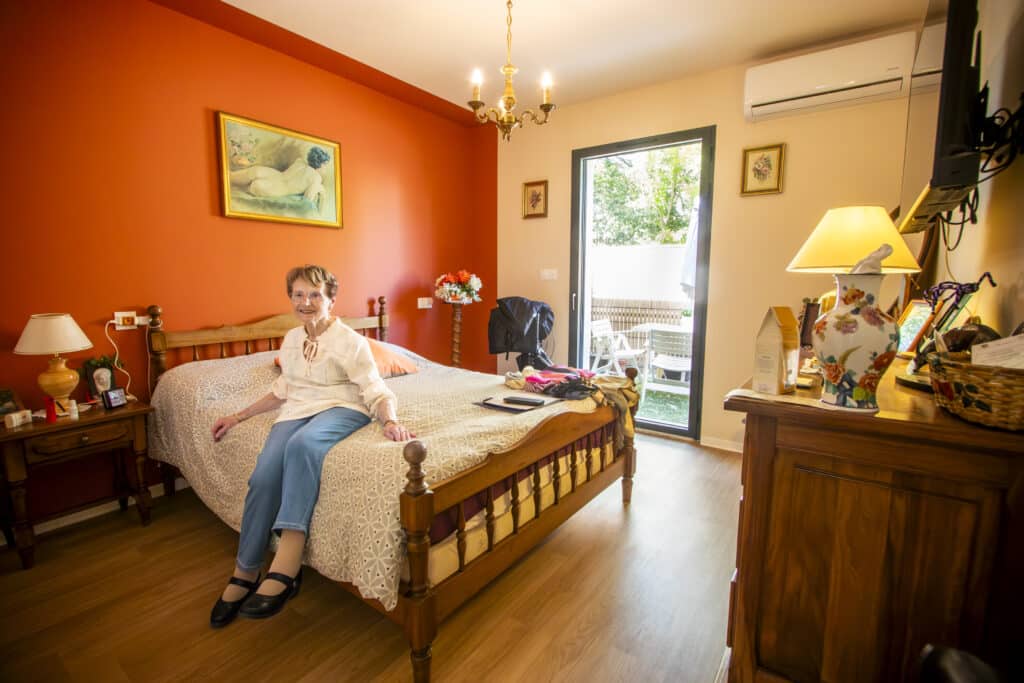 Femme âgée dans sa chambre au décor chaleureux et coloré Femme âgée souriante assise sur son lit dans une chambre décorée au mur orange avec accès terrasse.